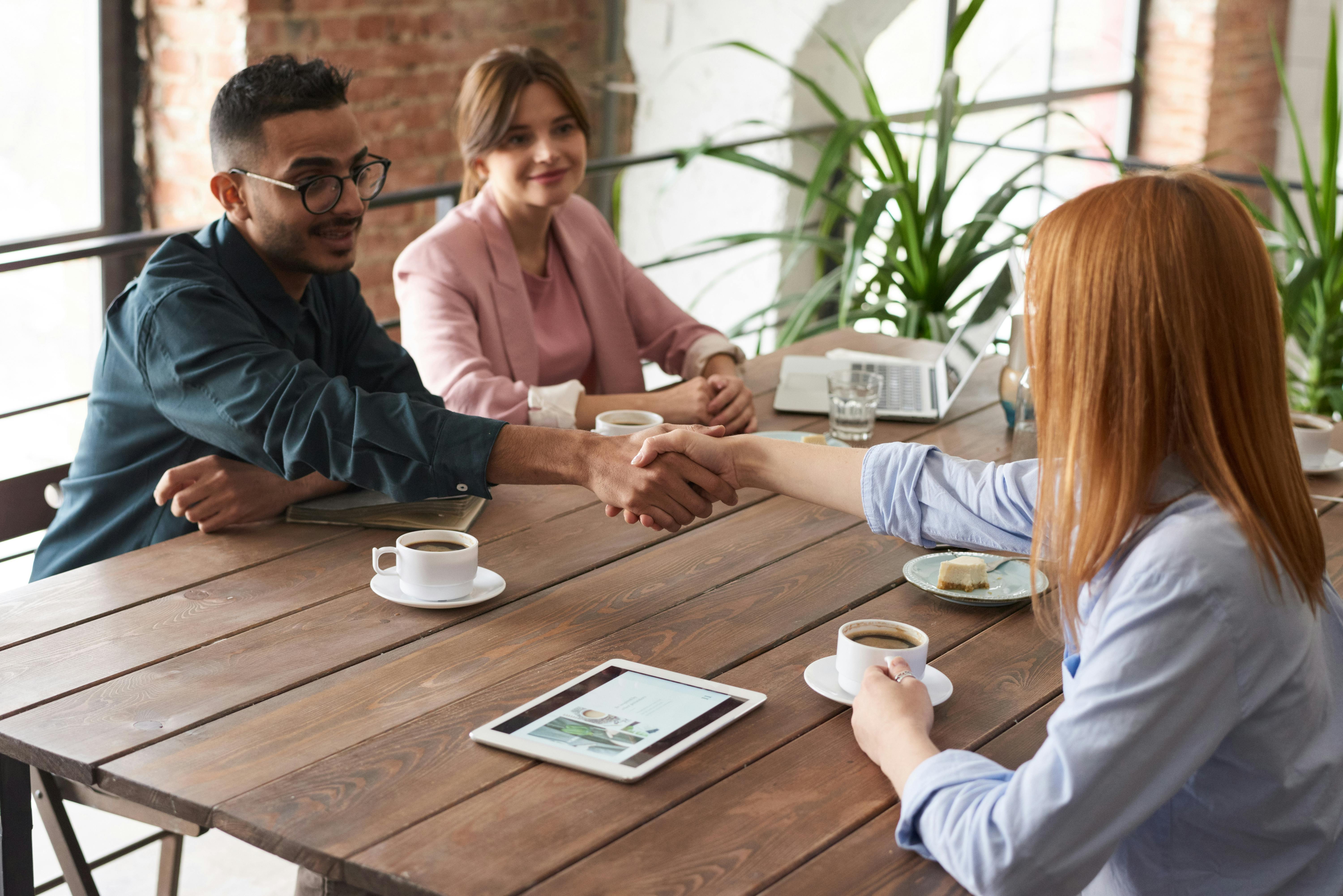 An image showing a group of young professionals sitting at a wooden table, with two people shaking hands in agreement.