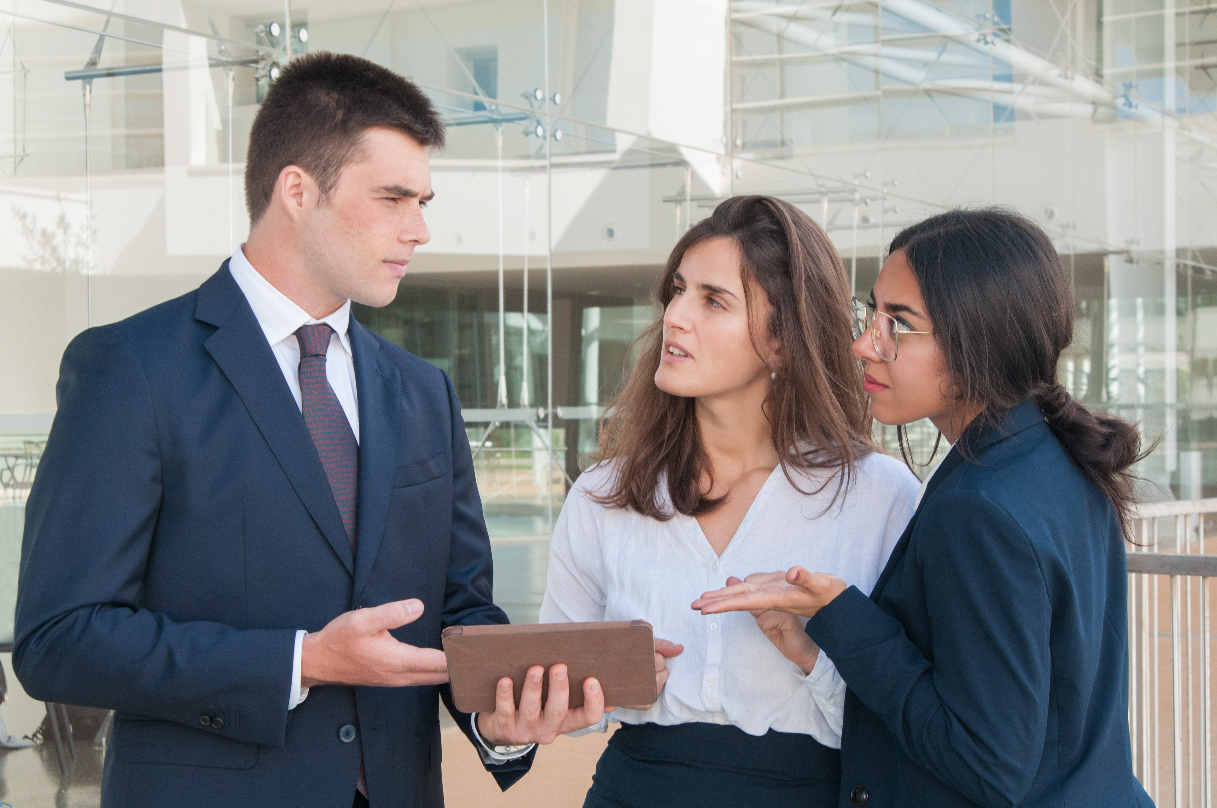 A businessman in a suit presents data on a tablet to two female colleagues during a serious discussion.